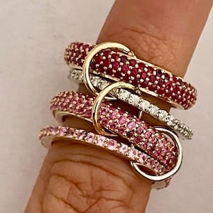 May include: An assortment of gemstone rings in shades of pink and red, with gold accents. The rings are encrusted with small stones and are stacked together on a finger, showcasing a variety of designs and colors.