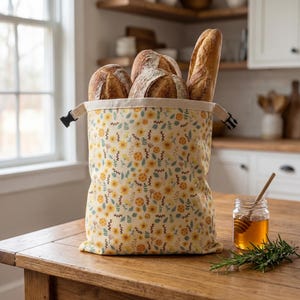 May include: A floral bread bag filled with various loaves of bread, including a baguette. The bag features a yellow and white floral pattern and a black buckle closure. A jar of honey and rosemary sprig are on the wooden table.