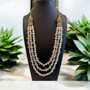 May include: A four-strand necklace with white oval beads and gold accents. The necklace has a green and gold pendant at each end.