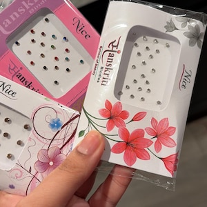 May include: Three packages of bindi, small decorative dots worn on the forehead. The packages are white, pink, and clear, with floral and decorative designs. Each package contains multiple small, colorful gems.