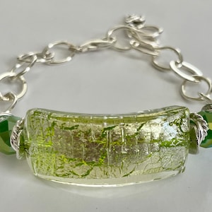 May include: A silver chain bracelet with a green glass bead and a silver charm. The glass bead has a textured surface and a metallic sheen.