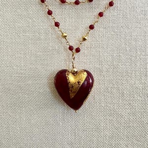 May include: A layered necklace featuring a heart-shaped pendant. The pendant is a deep red with gold leaf accents. The necklace has two strands of small red beads and gold-colored beads. The necklace is displayed on a neutral-colored fabric.