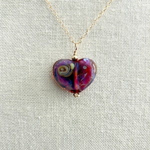 May include: A heart-shaped pendant necklace with a gold chain. The pendant is made of glass with a swirl design in shades of purple, red, and blue. The necklace is displayed on a neutral-colored fabric background.