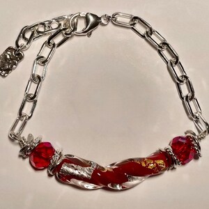 May include: A silver chain bracelet with a red and gold glass bead. The bead is twisted and has gold flecks throughout. The bracelet has a lobster clasp.