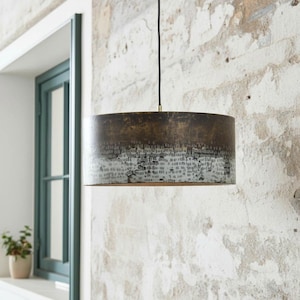 Può includere: Lampada a sospensione cilindrica con finitura bronzo anticato. La parte superiore è di un bronzo più scuro, che sfuma in un motivo più chiaro e strutturato verso il basso. Pende da un cavo nero.