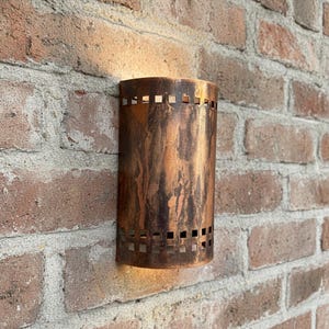 Puede incluir: Aplique de pared cilíndrico de color cobre con un acabado texturizado, montado en una pared de ladrillo rojo. El aplique tiene cortes cuadrados cerca de la parte superior e inferior, permitiendo que la luz brille. El aplique mide unos 25 cm de alto.