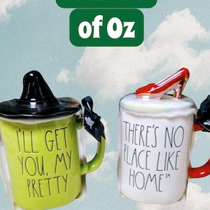 May include: Two ceramic mugs with Wizard of Oz themes. One is green with a black witch hat lid and the text "I'LL GET YOU, MY PRETTY." The other is white with a red shoe and the text "THERE'S NO PLACE LIKE HOME."