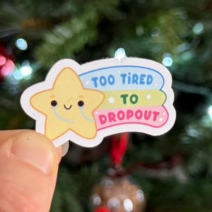 May include: A sticker featuring a yellow star with a smiling face and the text "TOO TIRED TO DROPOUT" in blue, yellow, and pink. The sticker is held in front of a Christmas tree with red ornaments.