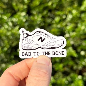 May include: A white sticker featuring a black and white illustration of a sneaker with the text "DAD TO THE BONE". The shoe design includes the letter "N" on the side. The sticker is held up against a blurred green background.