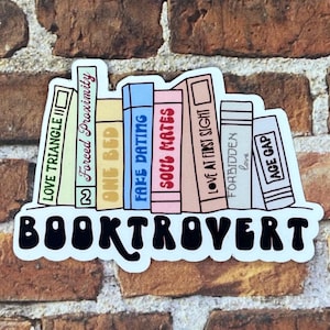 May include: A sticker featuring a stack of colorful books with titles like "Love Triangle II" and "Soul Mates." The word "BOOKTROVERT" is written in bold, black letters below the books. The sticker is on a brick background.