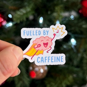May include: A sticker with a cartoon brain wearing a yellow cape and holding a star, with the text "FUELED BY CAFFEINE" in blue. The sticker is held in front of a Christmas tree with red ornaments and white lights.