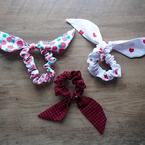 May include: Three fabric scrunchies with bow accents. One scrunchie features a white background with red strawberries, another has a white background with red hearts, and the third is red and black gingham. All are on a wooden surface.