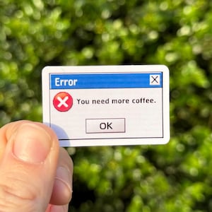 May include: A white sticker with a blue top bar that reads "Error." The sticker has a red "X" icon and the text "You need more coffee." Below is an "OK" button. The sticker is held in front of a green, blurred background.