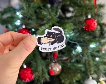 May include: A white sticker with a black cat design biting a hand. The text "TRUST NO CAT" is below the cat. The sticker is held in front of a Christmas tree decorated with red and silver ornaments.