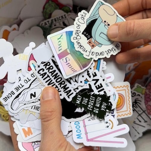 May include: A collection of various stickers with different designs and text. One sticker says "Bag of Cats" and another says "May the forest be with you". Other stickers have the words "You Smut", "Paranormal Cativity", and "Mood".