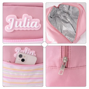 May include: Pink insulated lunch bag with the name "Julia" in white script. The bag has a zippered closure, a front pocket with a striped pattern, and a silver insulated interior. A smartphone is visible in the front pocket.