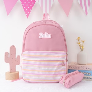May include: Pink backpack with a striped front pocket and the name "Julia" on the front. A matching pink pencil case sits next to the backpack. A pink cactus decoration and decorative flags are in the background.