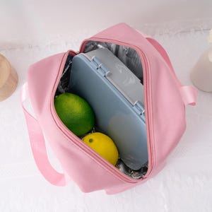 May include: A pink insulated lunch bag with a zipper closure, containing a blue lunch box, a green lime, and a yellow lemon. The bag has a silver insulated interior and a pink handle.