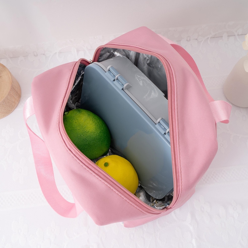 May include: A pink insulated lunch bag with a zipper closure, containing a blue lunch box, a green lime, and a yellow lemon. The bag has a silver insulated interior and a pink handle.