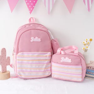 May include: A pink backpack and matching lunch bag set. The backpack and lunch bag have a striped front pocket and the name "Julia" on them. The set is displayed with pink decorative flags.