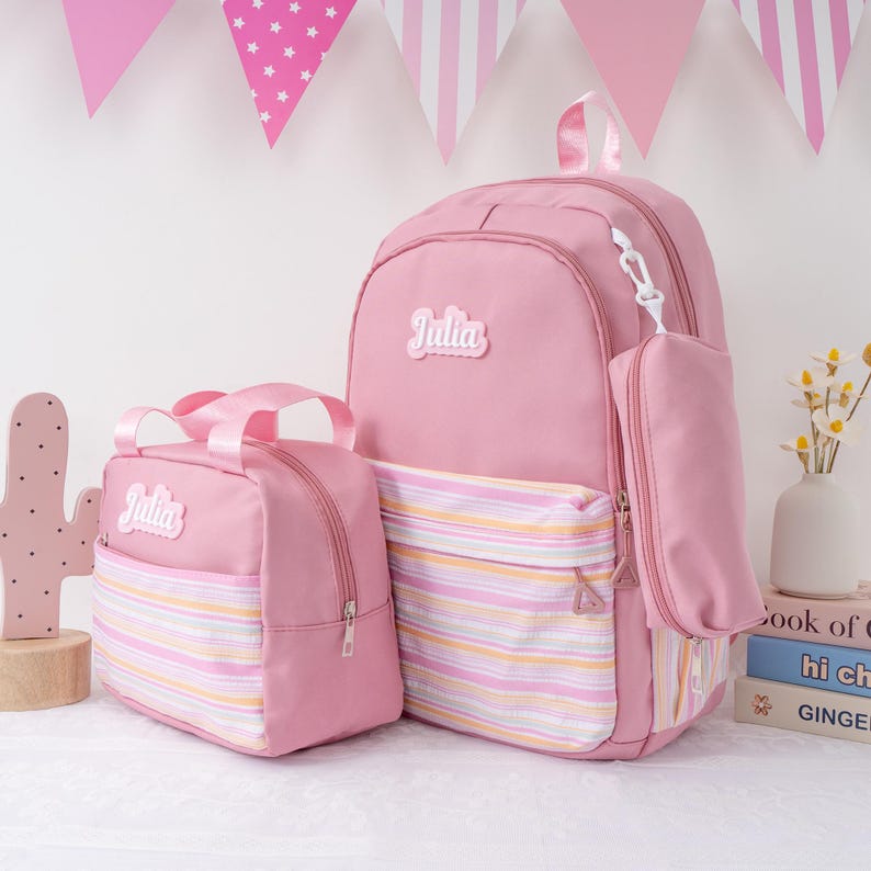 May include: Pink backpack and matching lunch bag set. The backpack has a front pocket with a striped design and the name "Julia" on the top. The lunch bag has a matching striped design and the name "Julia".