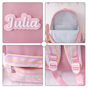 May include: Pink backpack with a striped front pocket and adjustable straps. The backpack has the name "Julia" in white script on the front. The interior is light gray.