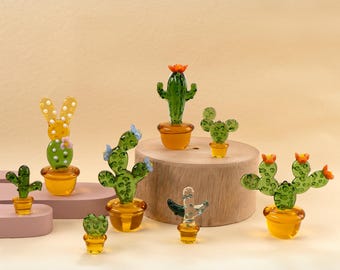 May include: A collection of glass cactus figurines in various shapes and sizes. The cacti are green with orange, yellow, and blue accents, and sit in yellow-orange pots. Some have flower details. The figurines are displayed on a light brown surface.