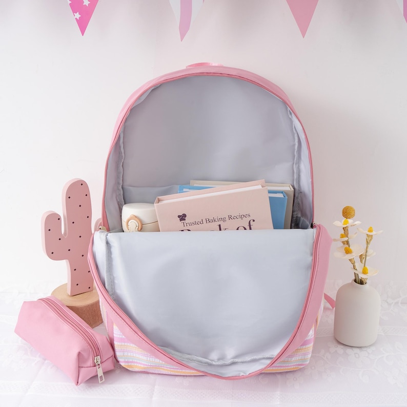 May include: Pink backpack with a matching pencil case, open to reveal books and a water bottle. The backpack has a striped design at the bottom and is set against a white background with decorative elements.