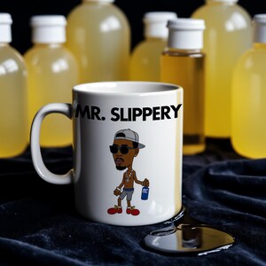 May include: White ceramic mug with a black cartoon illustration of a person wearing sunglasses and a hat. The mug has the text "MR. SLIPPERY" printed on it. The mug is sitting on a dark blue surface with a black liquid spill.