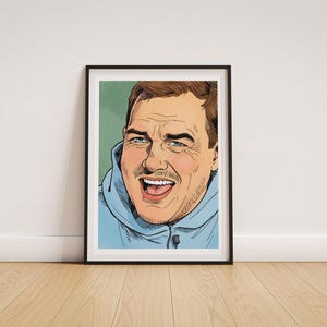 May include: A framed art print featuring a comic book style illustration of a person with brown hair and a light mustache, wearing a light blue hooded sweatshirt. The person has a wide, laughing expression with visible teeth against a green background.