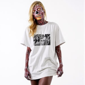 Op de afbeelding: Een wit T-shirt met een zwart-witfoto van een vrouw en twee mannen. De foto is gedrukt op de voorkant van het shirt en de tekst "BETRAY" is gedrukt boven de foto.