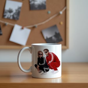 May include: White ceramic mug featuring a color image of two people in tracksuits and sunglasses. One person wears a red tracksuit with white stripes, the other wears a black tracksuit with white stripes. The mug has a curved handle.