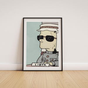 May include: A framed illustration featuring a cartoon character wearing sunglasses and a traditional head covering, dressed in a grey military-style uniform. The character sits behind a desk with a nameplate that clearly displays the text LIBYA.