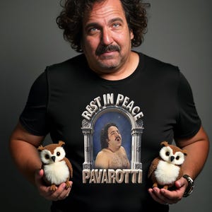 May include: A man wearing a black t-shirt with the text "REST IN PEACE PAVAROTTI" and a portrait of a man. He is holding two small brown and white owl plush toys.
