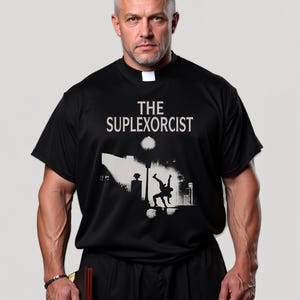 Könnte beinhalten: Schwarzes T-Shirt mit dem weißen Text "THE SUPLEXORCIST" und einer Grafik von zwei Figuren, die unter einer Straßenlaterne ringen. Das T-Shirt wird von einer Person mit weißem Priesterkragen getragen.