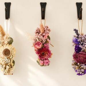 May include: Three hanging floral arrangements made of dried flowers. The left bundle features cream and beige tones, the centre bundle displays shades of pink and magenta, and the right bundle showcases deep purple and violet hues, all suspended from black hooks.