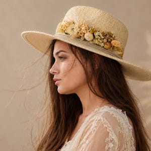 Könnte beinhalten: Ein Strohhut mit breiter Krempe, verziert mit getrockneten Blumen in Creme- und Gelbtönen. Der Hut hat ein helles Band. Die Person, die den Hut trägt, hat lange, braune Haare. Der Stil ist bohème und natürlich, mit floralen Akzenten.