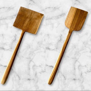 May include: Two wooden spatulas with long handles and rectangular and rounded heads. The spatulas are made of light brown wood and are set against a white and grey marble background. The wood grain is visible.