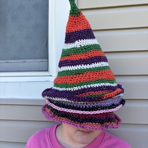 May include: A tall, pointed crochet hat featuring horizontal stripes in orange, purple, green, and white. The hat has a wide, slightly floppy brim and a textured, yarn-based construction with a metallic shimmer throughout the fabric.