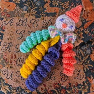May include: A handmade crochet clown doll with a white head, orange cone hat, and colorful spiral body. The clown's face has blue star eyes and a small orange nose. The body is made of turquoise, yellow, and purple crochet.