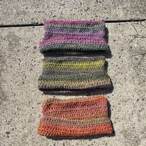 May include: Three handmade crochet beanies arranged vertically. The top beanie features variegated purple and green yarn. The middle beanie displays variegated olive green and grey tones. The bottom beanie is crafted from variegated orange and pink yarn.