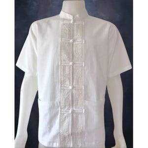 Puede incluir: Camisa blanca de manga corta con cuello mandarín y cierres decorativos de nudo en la parte delantera.
