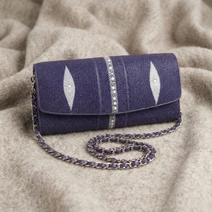 May include: A deep purple clutch with a silver chain strap. The clutch features a textured surface with white diamond-shaped accents and a central silver beaded stripe. The chain is a matching purple and silver.