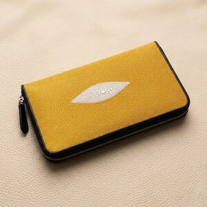 May include: A yellow leather wallet with a white oval design in the centre. The wallet has a black zip and trim. The texture of the leather is visible. The wallet is rectangular and appears to be closed.