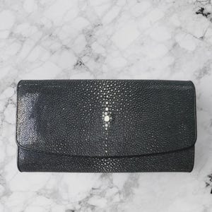 May include: A dark grey wallet with a textured surface and a flap closure. The wallet features a pattern of small, light-coloured dots and a central, larger, white dot. The wallet is rectangular and appears to be made of leather.