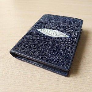 Könnte beinhalten: Ein marineblaues Lederportemonnaie mit einem weißen Stachelrochen-Design. Das Portemonnaie ist rechteckig und hat eine strukturierte Oberfläche. Das Stachelrochen-Design befindet sich in der Mitte.