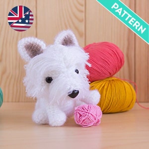 Puede incluir: Juguete de peluche de perro blanco tejido a crochet con orejas marrones, sentado sobre una superficie de madera con ovillos de lana en rosa, amarillo y rojo.