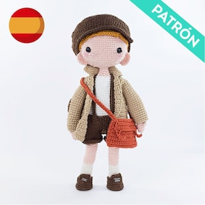 May include: Crocheted doll with brown hair, wearing a brown hat, a beige jacket, brown shorts, and a red bag. The doll is standing on a white background.