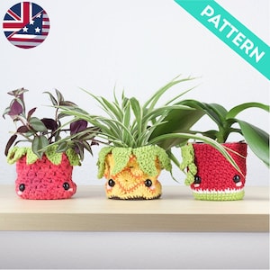 Könnte beinhalten: Drei gehäkelte Pflanzentopf in Form von Früchten. Die Töpfe sind rot, gelb und grün und haben süße Gesichter. Die Töpfe sind mit Pflanzen gefüllt.
