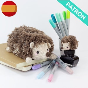 May include: A brown crocheted hedgehog with a white face and black eyes. The hedgehog is lying on a stack of notebooks and there are several colored markers next to it. There is a smaller crocheted hedgehog on top of a black ink bottle.
